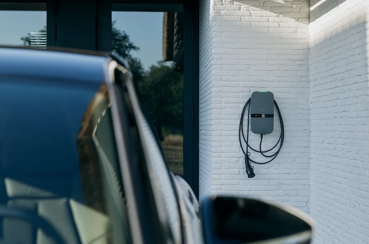 Peblar Home laadpaal met vaste laadkabel aan een witte gevel, naast een elektrische auto op de oprit