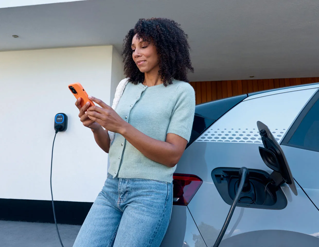 Compacte Wallbox Pulsar Max laadpaal aan een lichte gevel, terwijl een elektrische auto ernaast laadt en de app wordt bediend